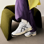 Model seated on olive-green suede chair wearing white and silver-grey ASICS sneakers and purple trousers